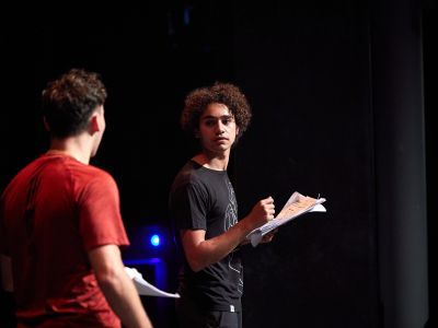 Two men on a stage turning towards each other, both are holding a script